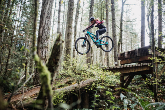 An adult woman on a mountain bike flies through the air off of a jump on a Pacific Northwest forest mountain bike trail. Shot in Issaquah, Washington, USA.