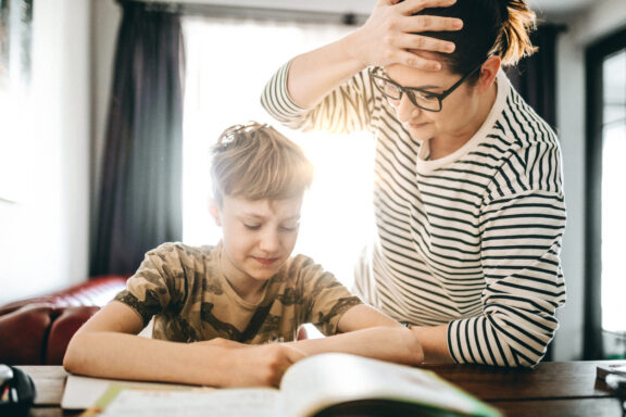 Having problems in finishing homework. Sad teenage boy doing homework at home and his upset mother is standing next to him