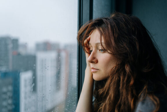 Young woman feeling sad and pensive, experiencing autumn depression while looking out a rainy city window, pondering loneliness and mental health challenges during a melancholic season