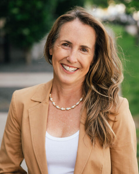 Smiling woman with long light brown hair wearing a tan blazer and white top, standing outdoors in a park.