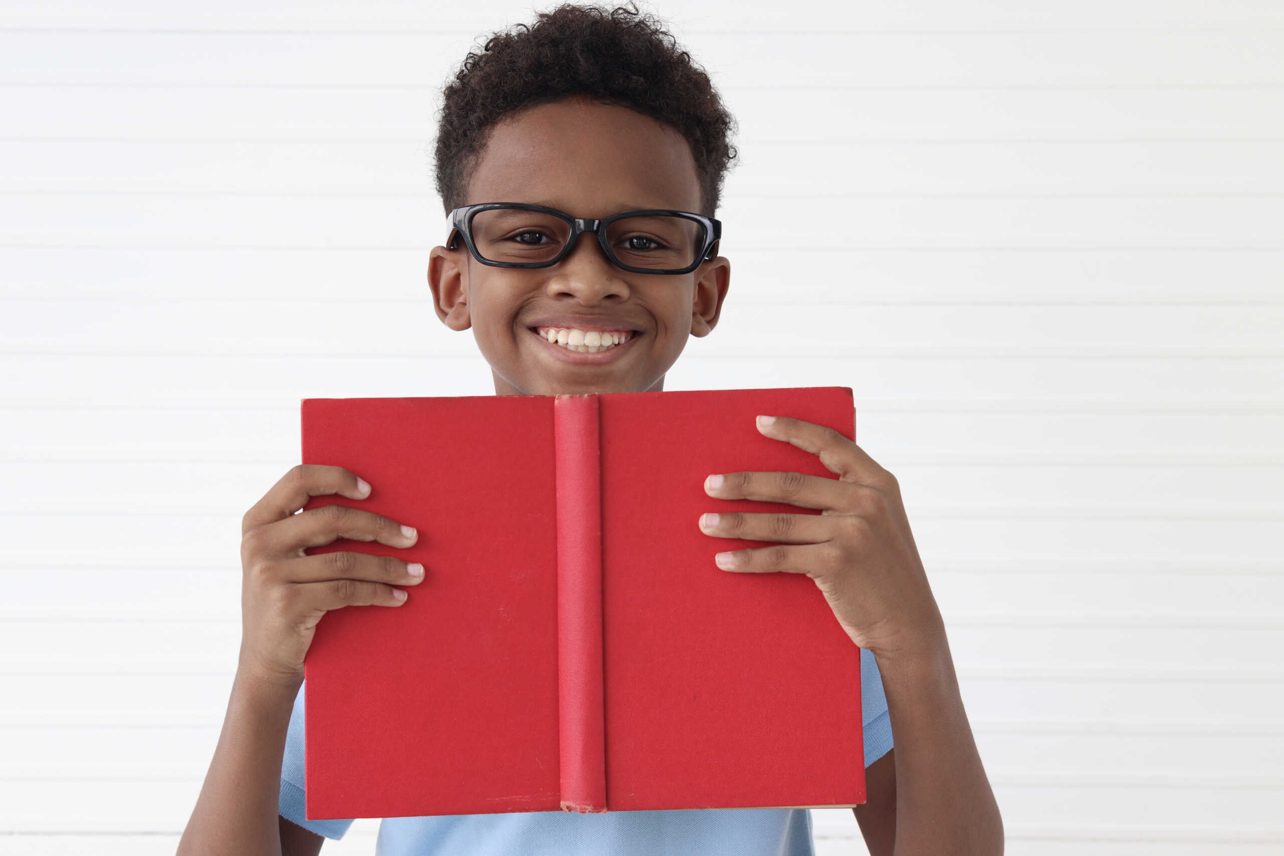 Smiling child with glasses holds a red book in front of a light, striped background.