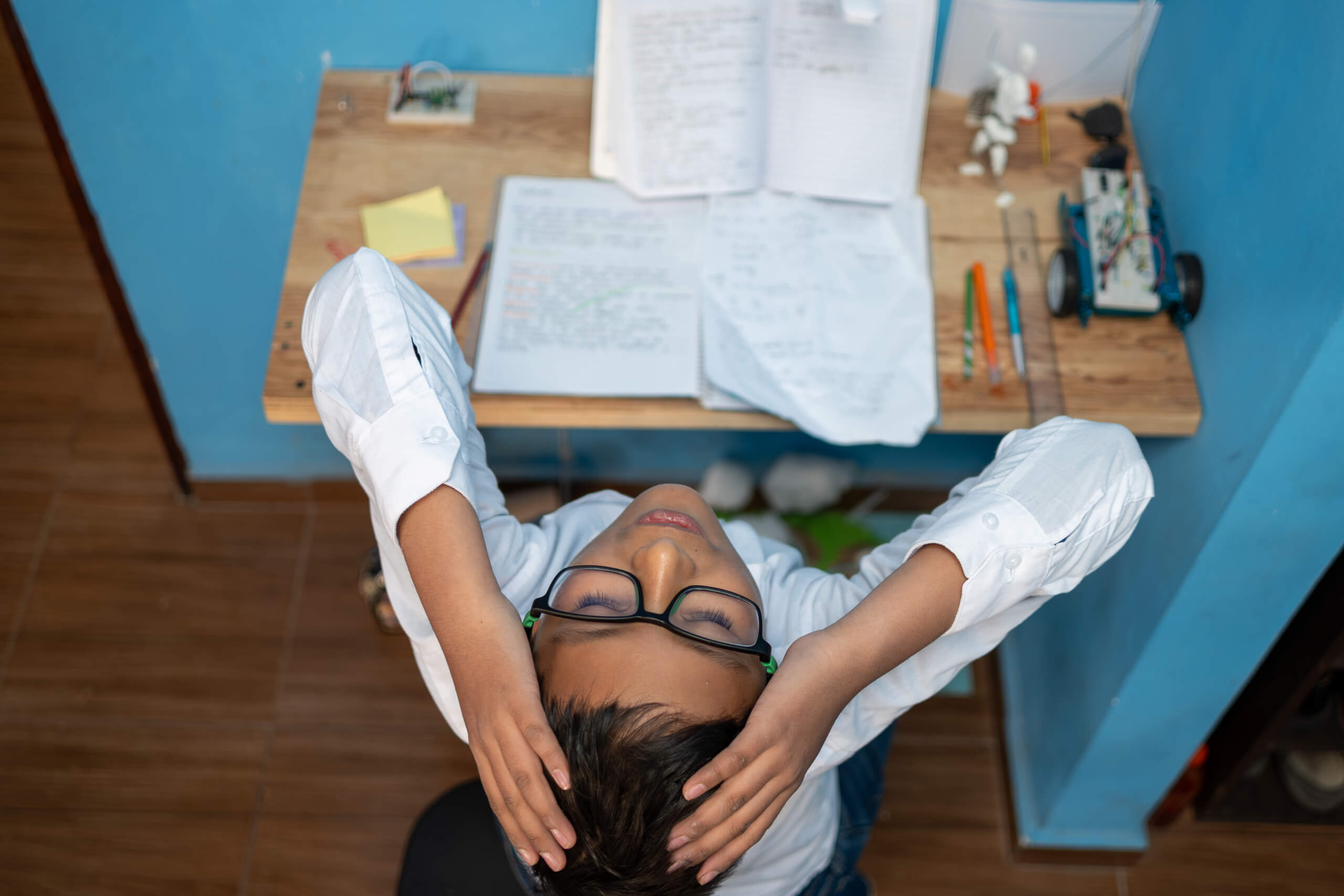 Top-down view of a person in a white lab coat with glasses, sitting at a cluttered desk with notebooks and a small robot on the right.